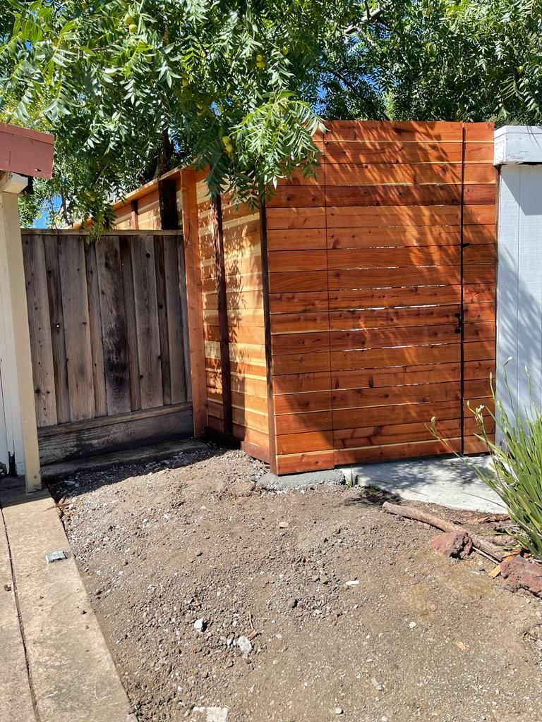 Wooden shed with horizontal planks, next to a weathered vertical-plank fence, on a gravel surface.