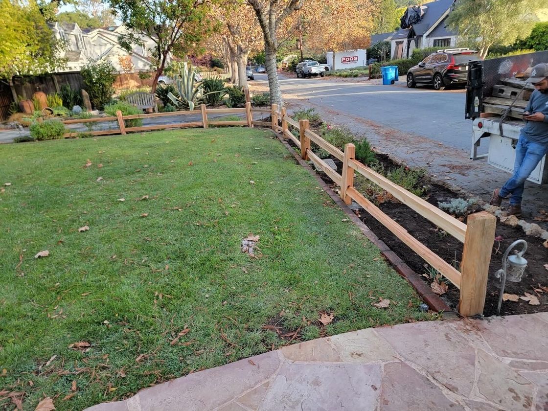 Wooden split-rail fence in a grassy yard, along a street. Man unloading supplies from a truck.