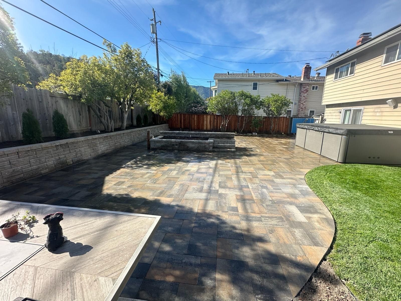 Backyard with stone patio, hot tub, grass, and retaining walls, under a blue sky.