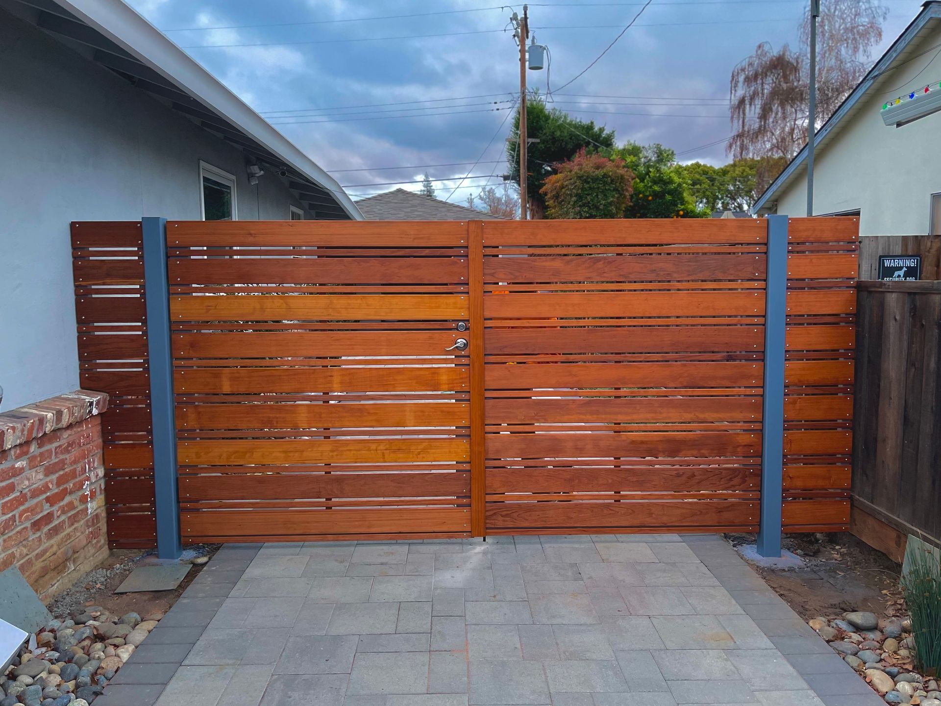Wooden slatted driveway gate with blue posts between two houses, viewed from a paved courtyard