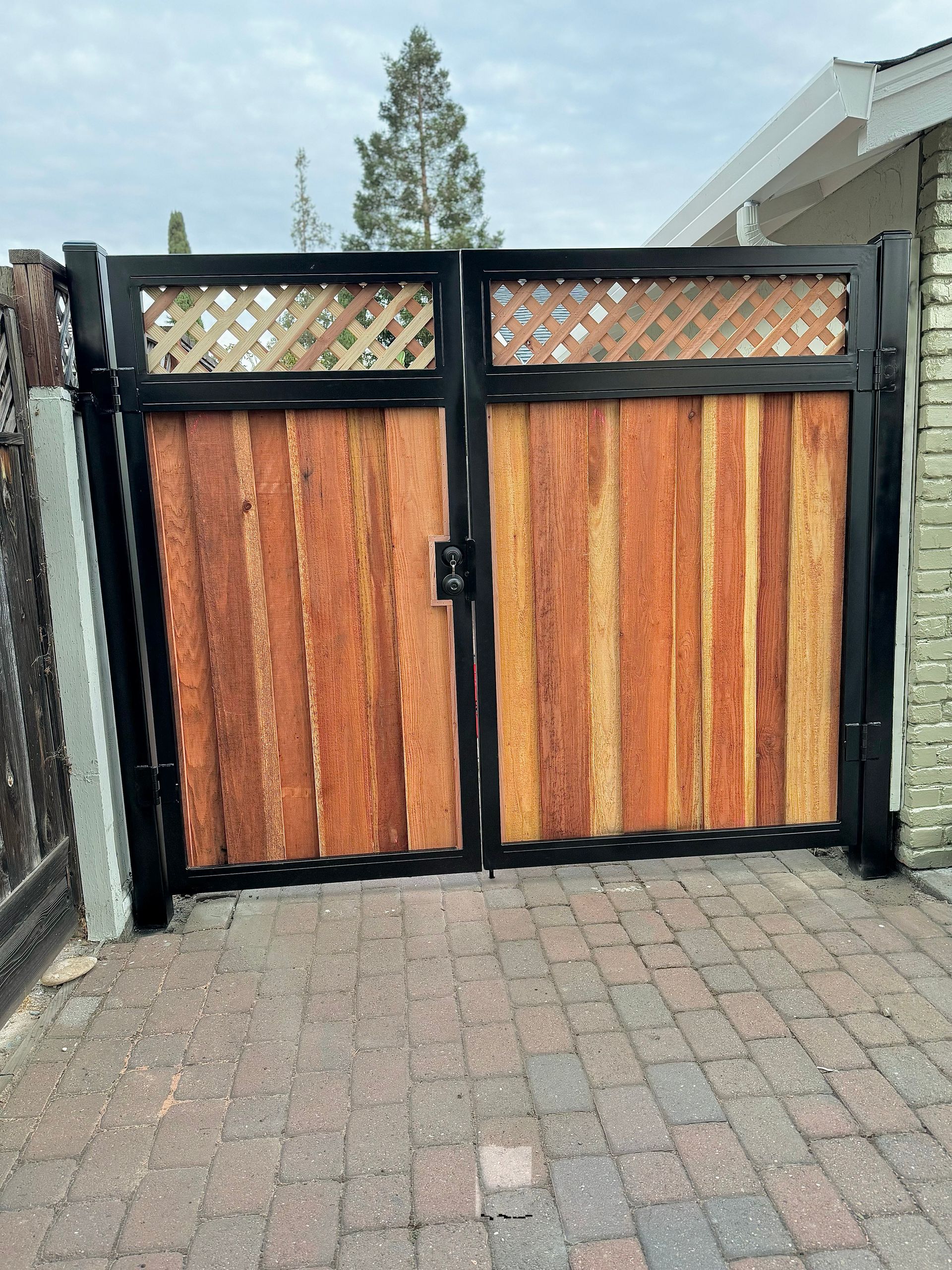 Black metal and wood gate with lattice top, set in a brick driveway.