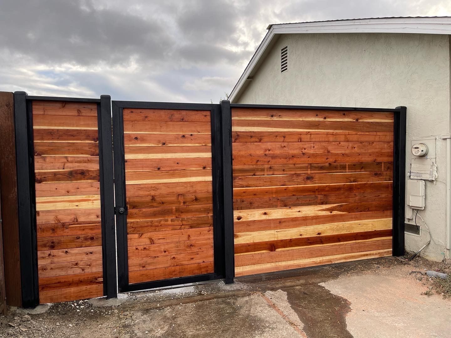 Wooden gate with black frame attached to a building, against a cloudy sky.