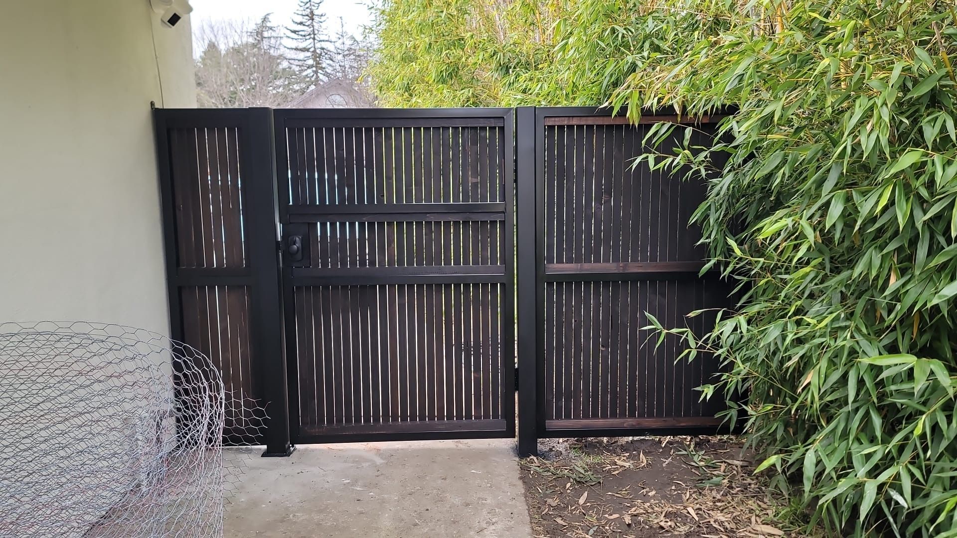 Black wooden gate with woven reed panels, set against a concrete surface with greenery.