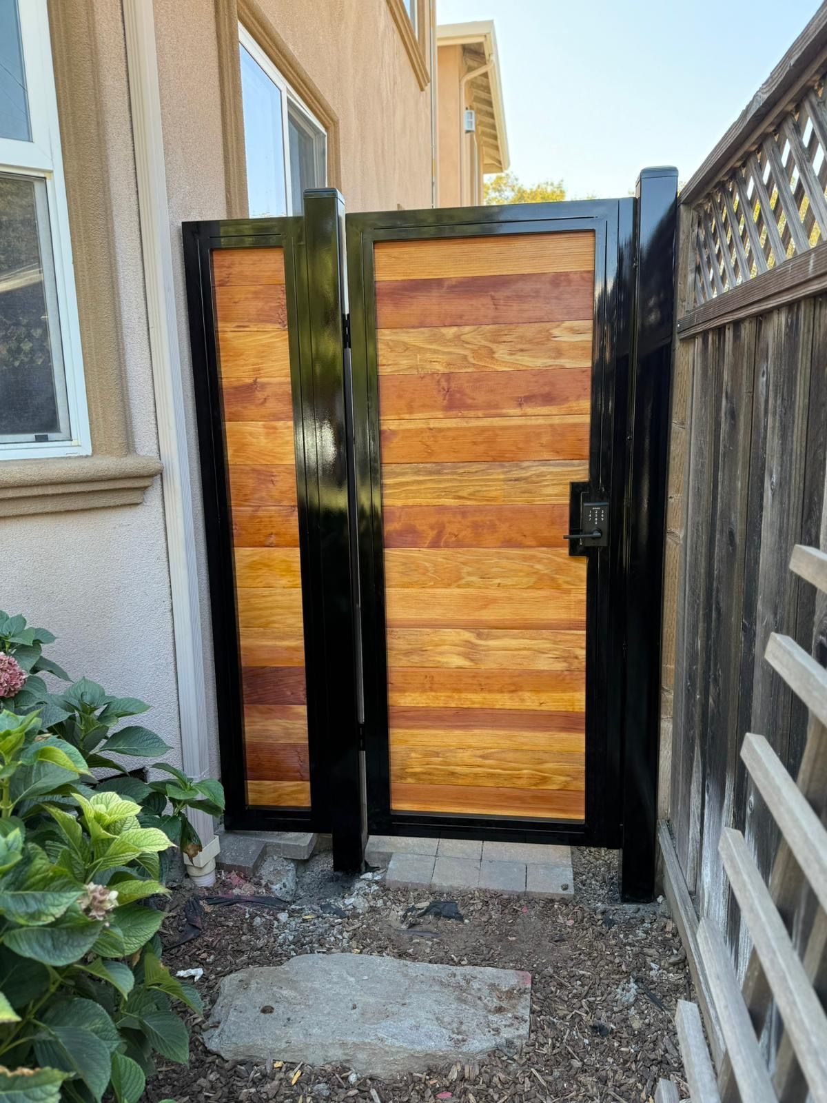 A modern gate with horizontal wood planks framed in black metal, installed between a house wall and a wooden fence.