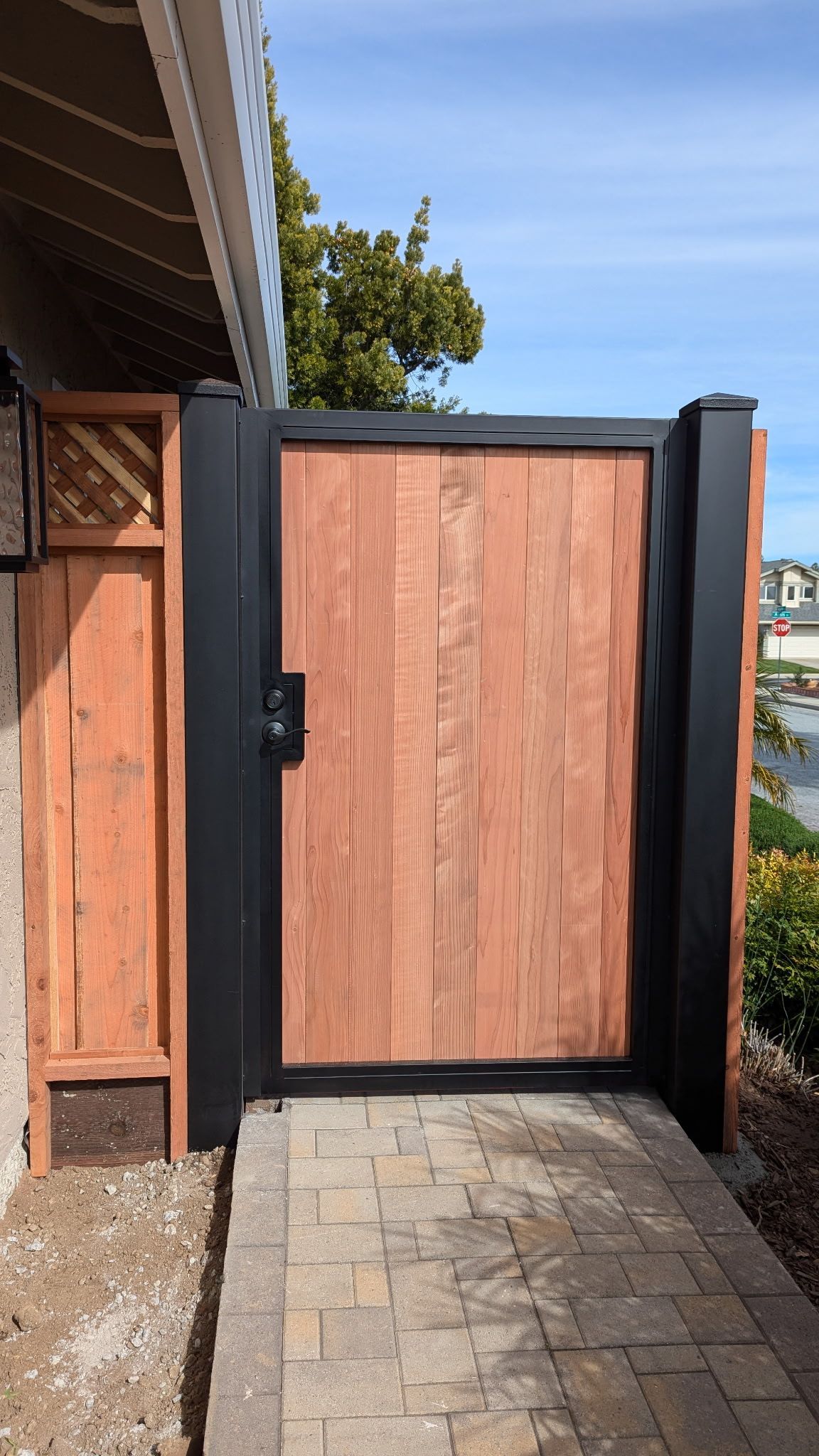 A modern wooden gate with a black metal frame stands at the end of a stone paver walkway next to a wooden fence.