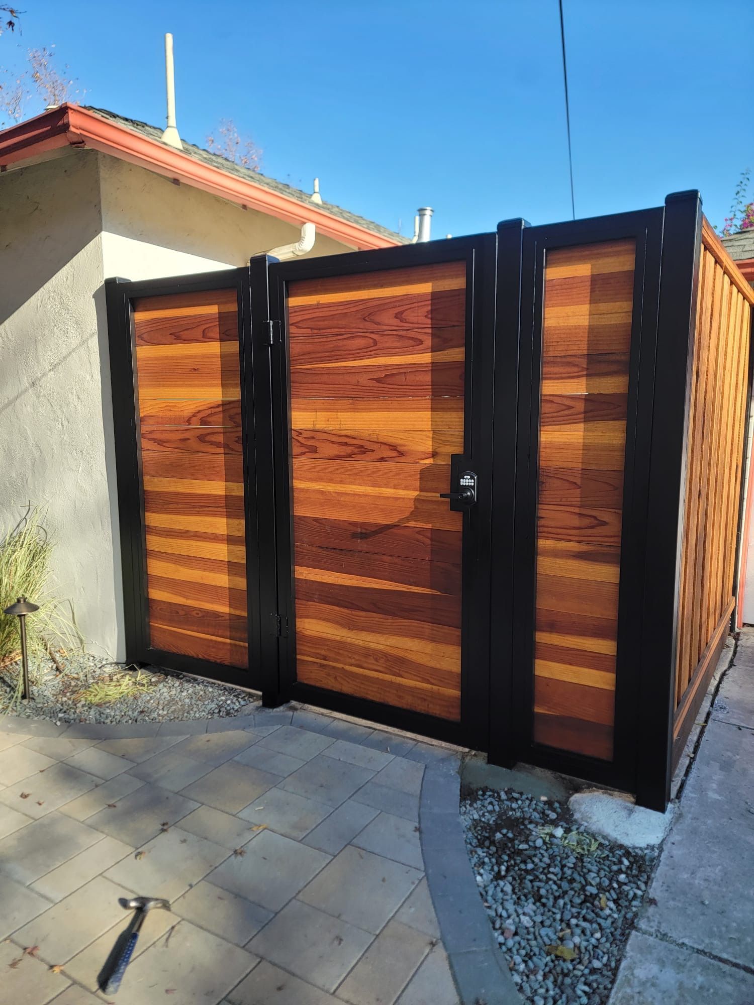 Wooden double gate with black metal frame; set in a paved area, connecting to a home.