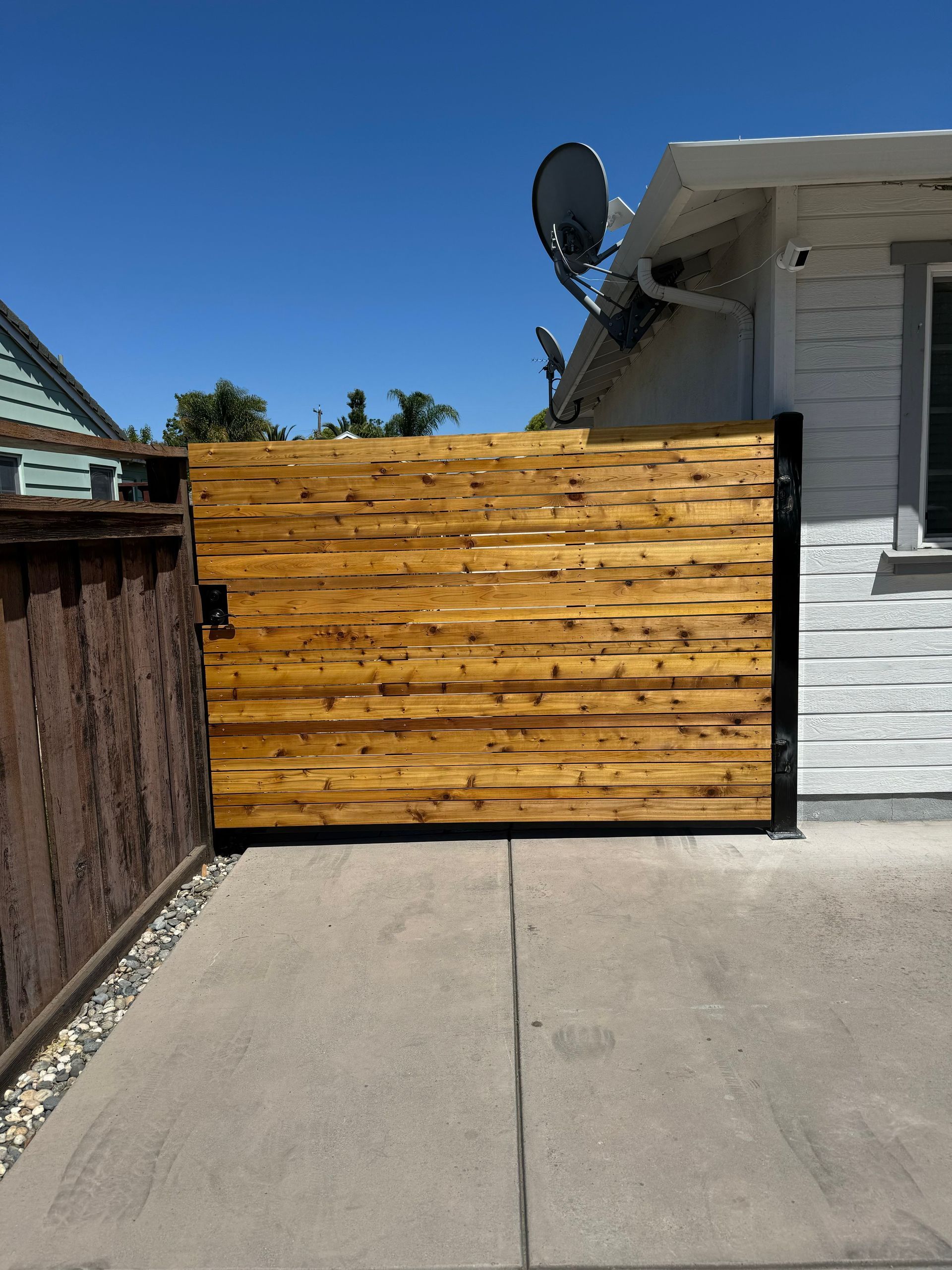 A new, horizontally-slatted wood driveway gate stands between a house and a wooden fence on a concrete driveway.
