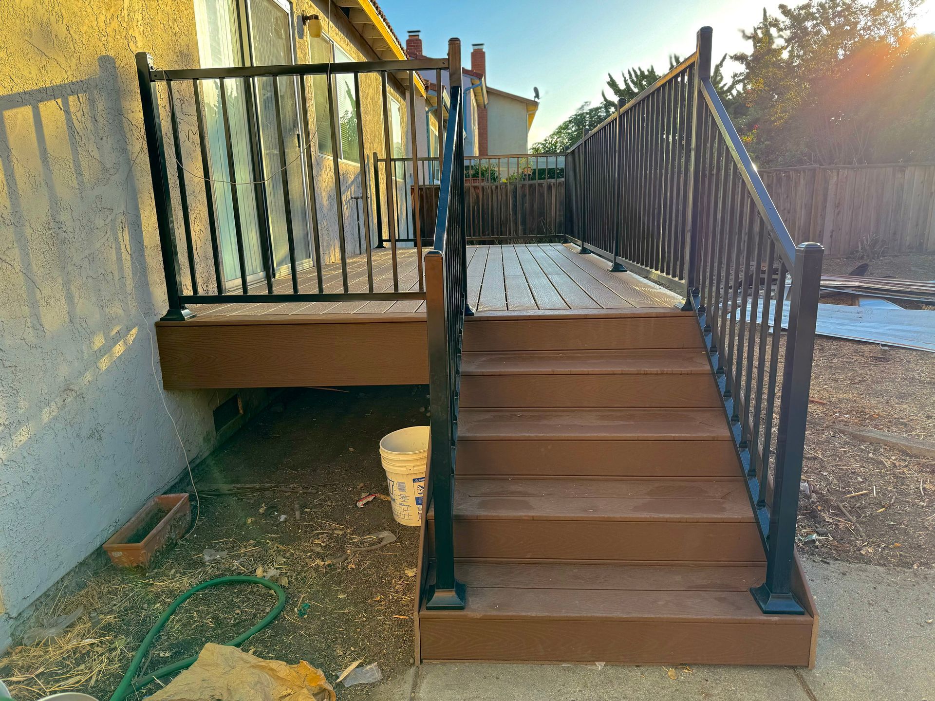 Brown wooden deck with stairs and black metal railings against a house in a sunny backyard.