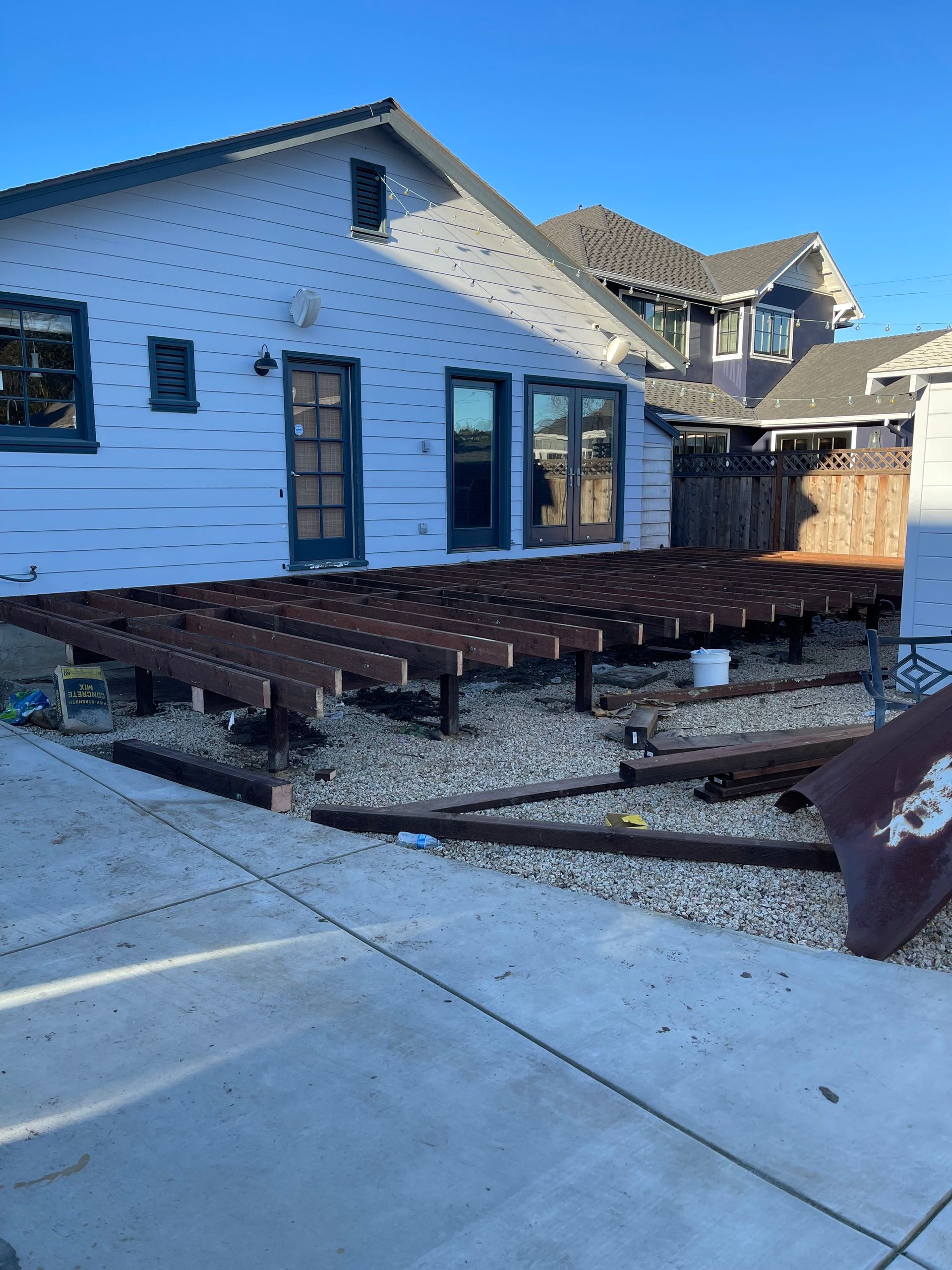 A white wooden porch walkway with a railing, bordered by green bushes on a sunny day.