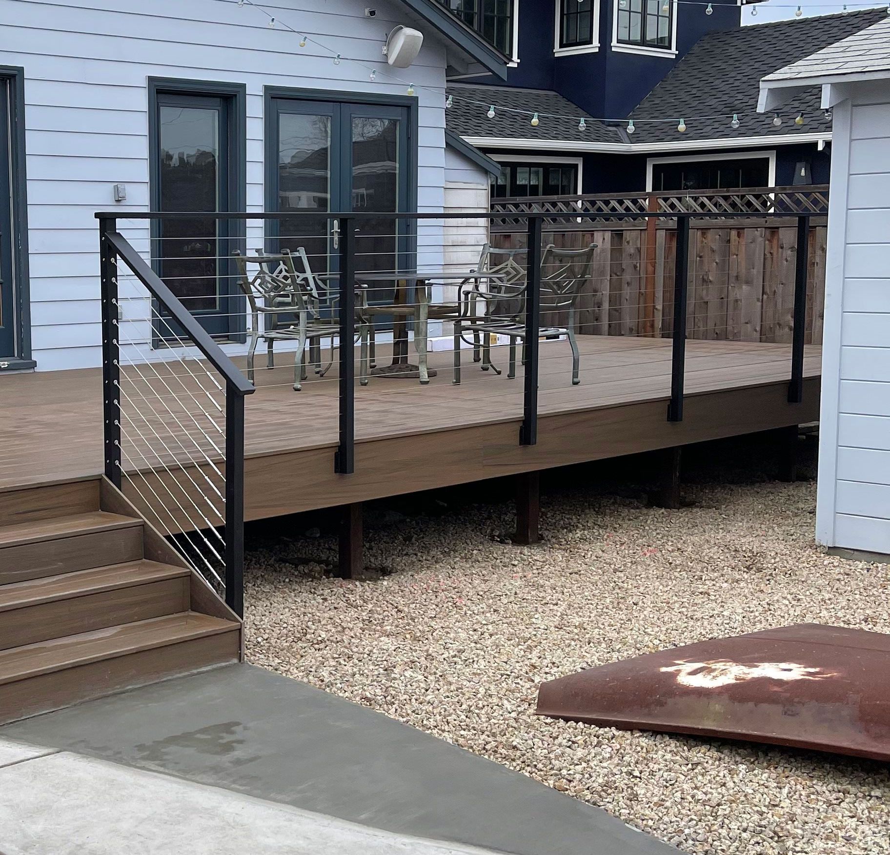 A raised brown deck with a modern black railing and cable infill, connected to a light blue house by a set of steps.