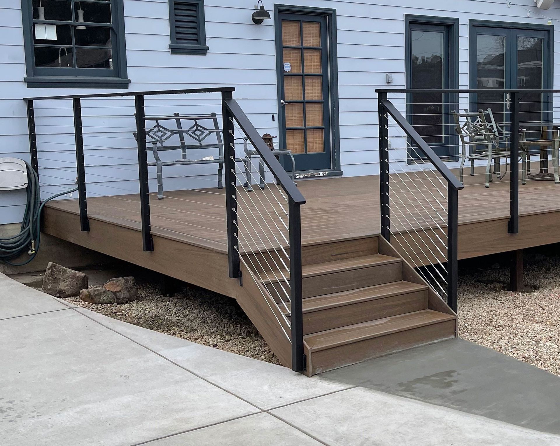 A wooden deck with a modern cable railing and short steps, attached to a light blue building with a concrete patio.