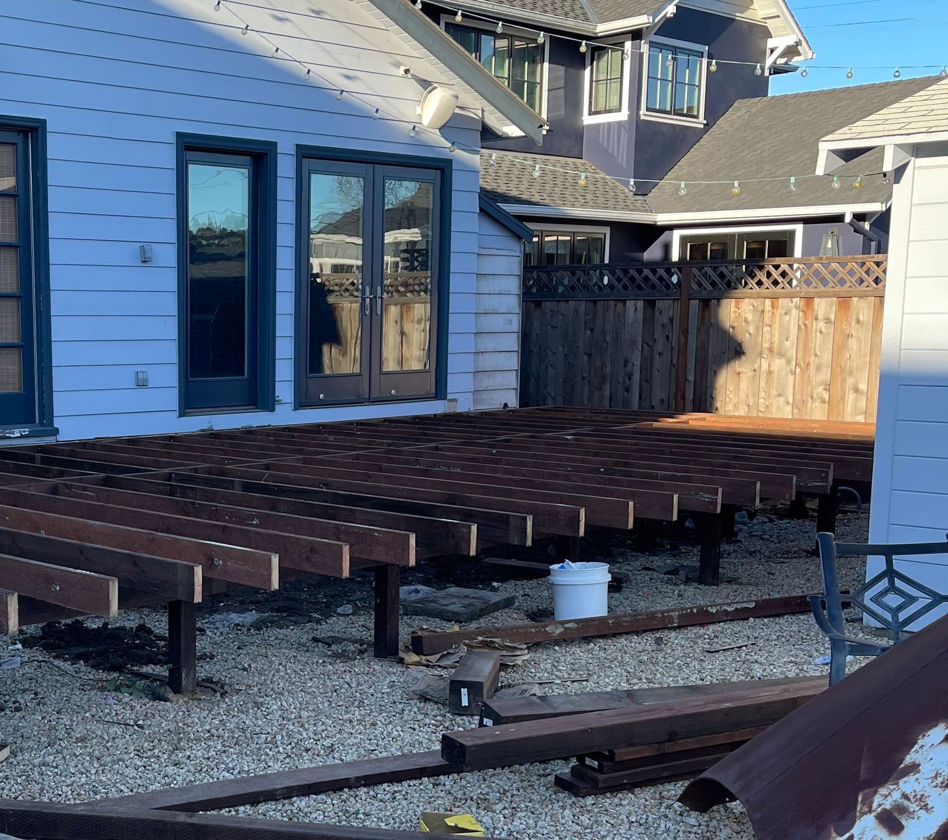 A residential outdoor deck under construction, featuring exposed wooden joists supported by posts over a gravel yard.