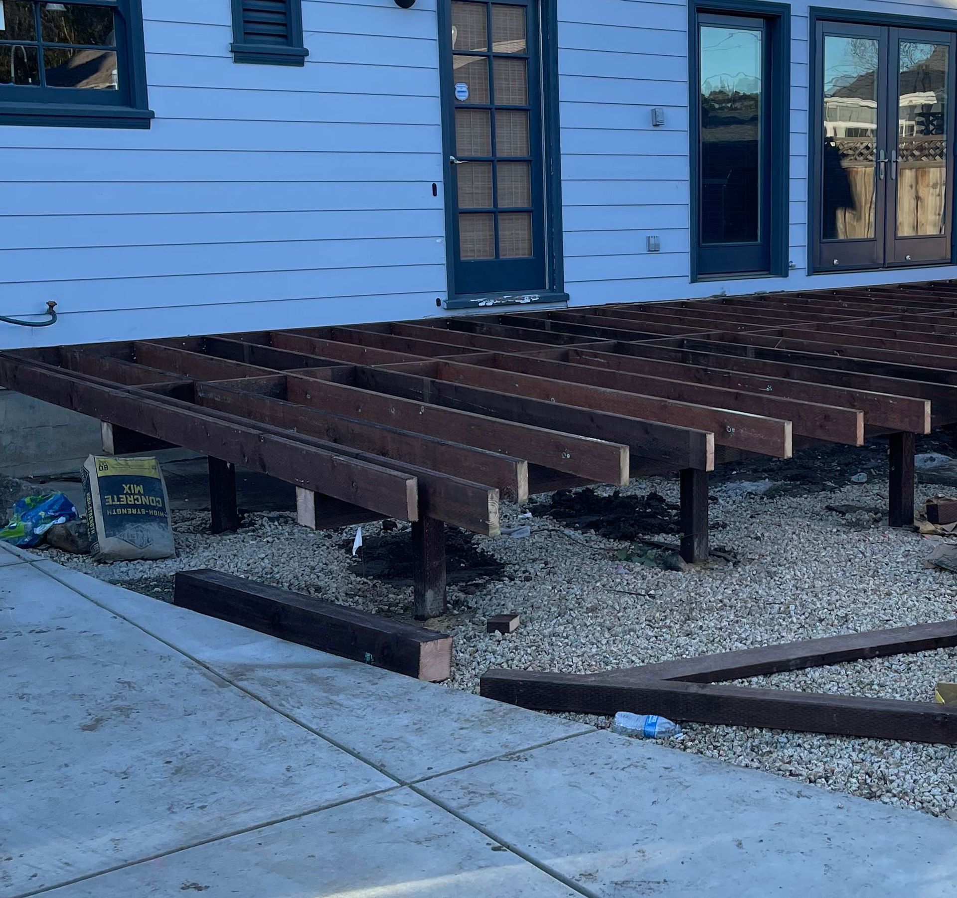 A wooden deck frame under construction against the side of a light blue house, situated over a gravel and concrete patio.