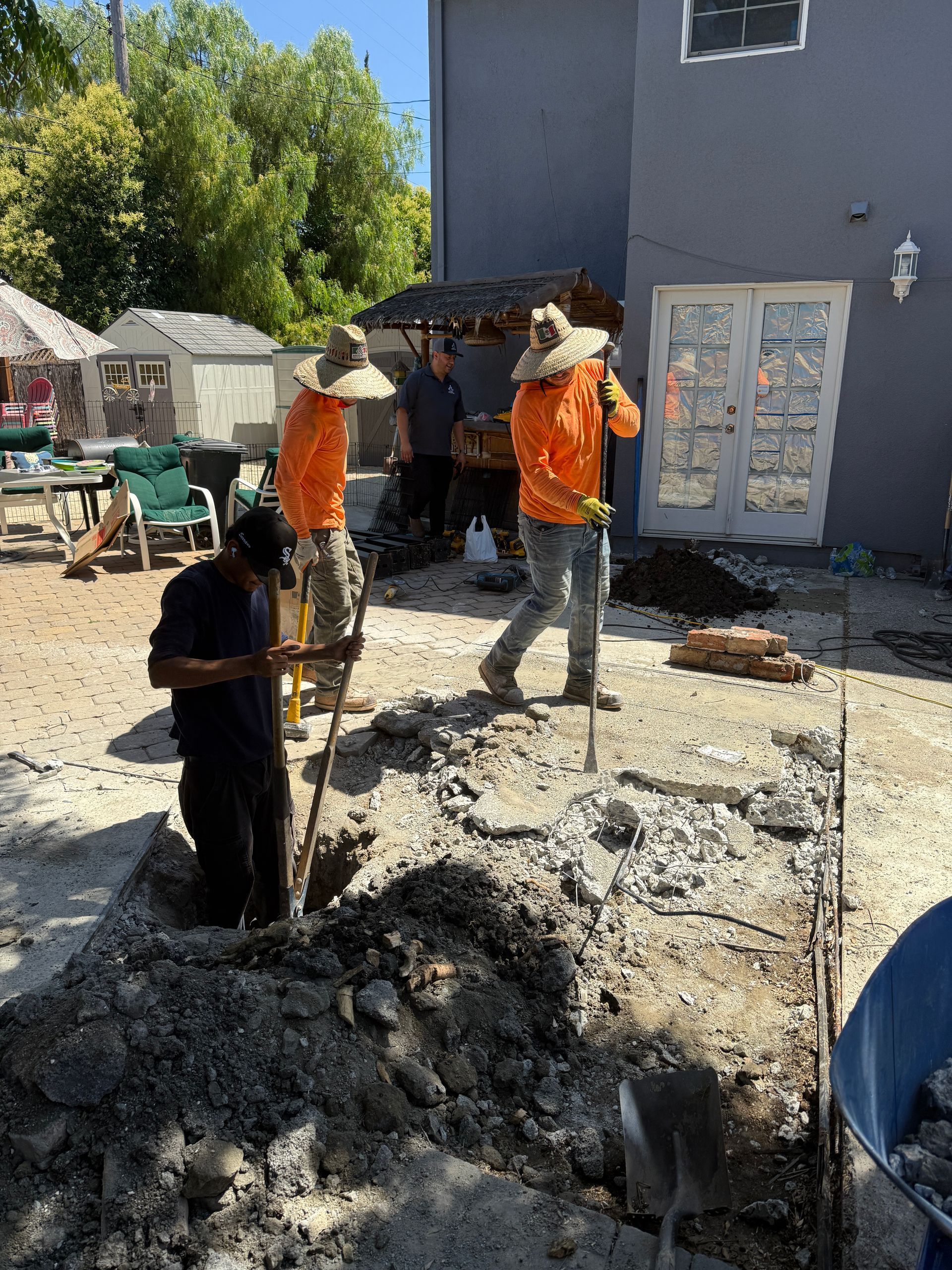 Construction workers digging in a backyard, wearing orange shirts and straw hats under a sunny sky.