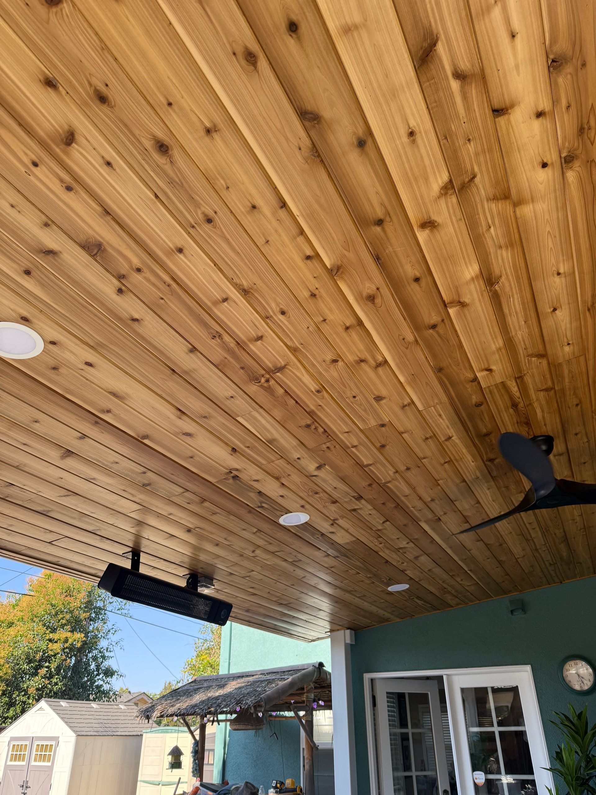 Wooden ceiling of a patio with recessed lights, a fan, and a heating element.
