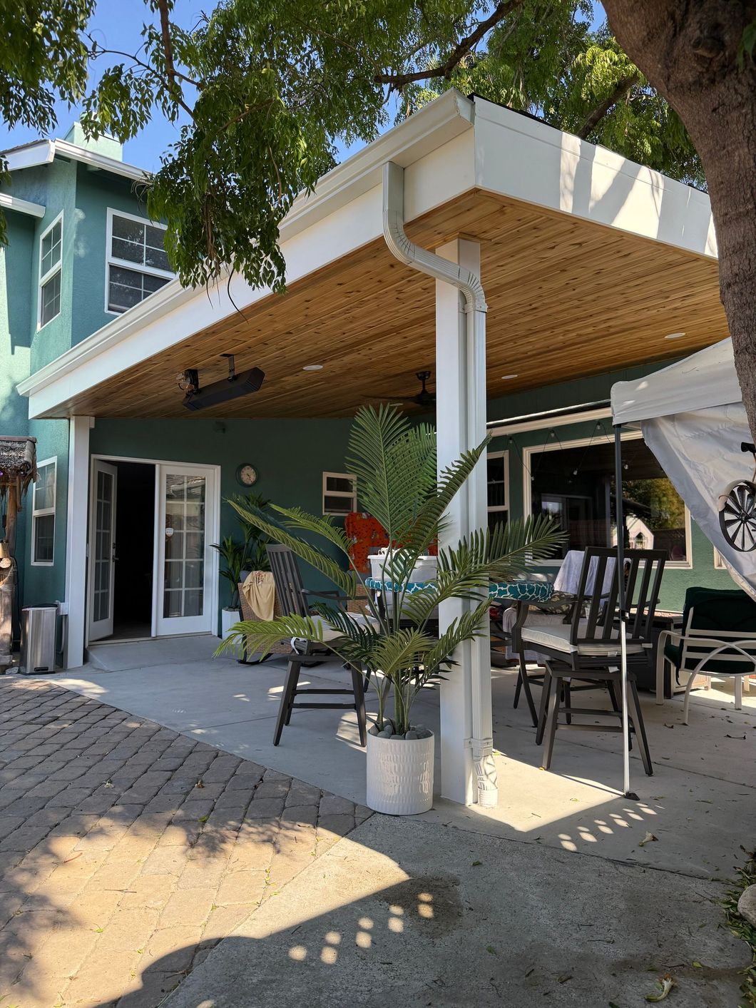 Backyard patio with green house, white pillars, dining table, and wicker ceiling.