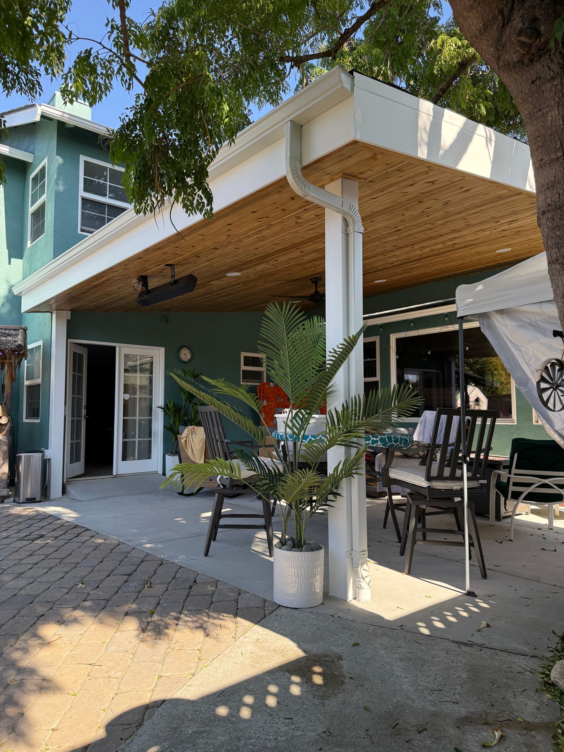 Backyard patio with green house, white pillars, dining table, and wicker ceiling.