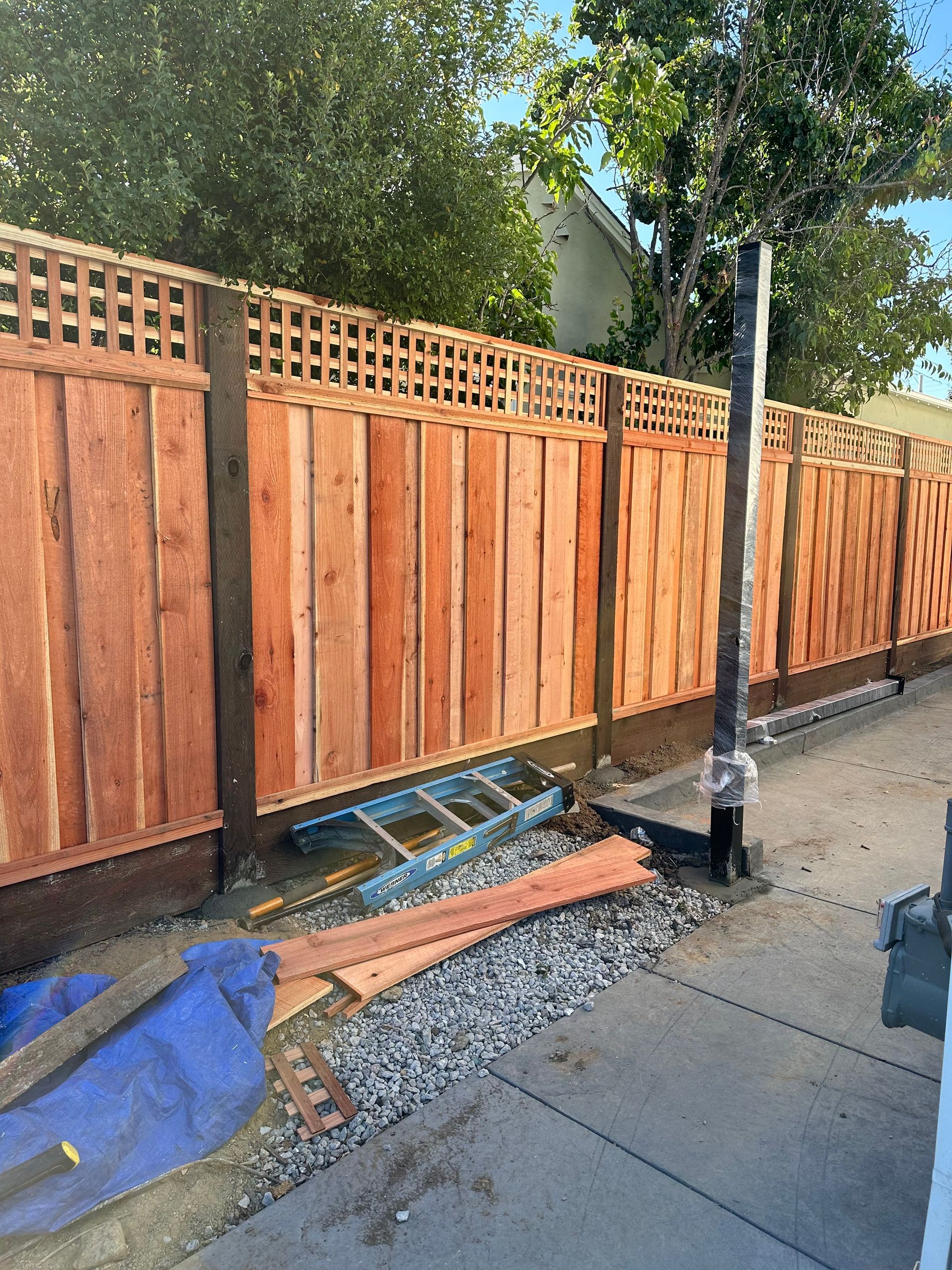 A new cedar wood fence with a lattice top border stands along a sidewalk, with construction materials on the ground.