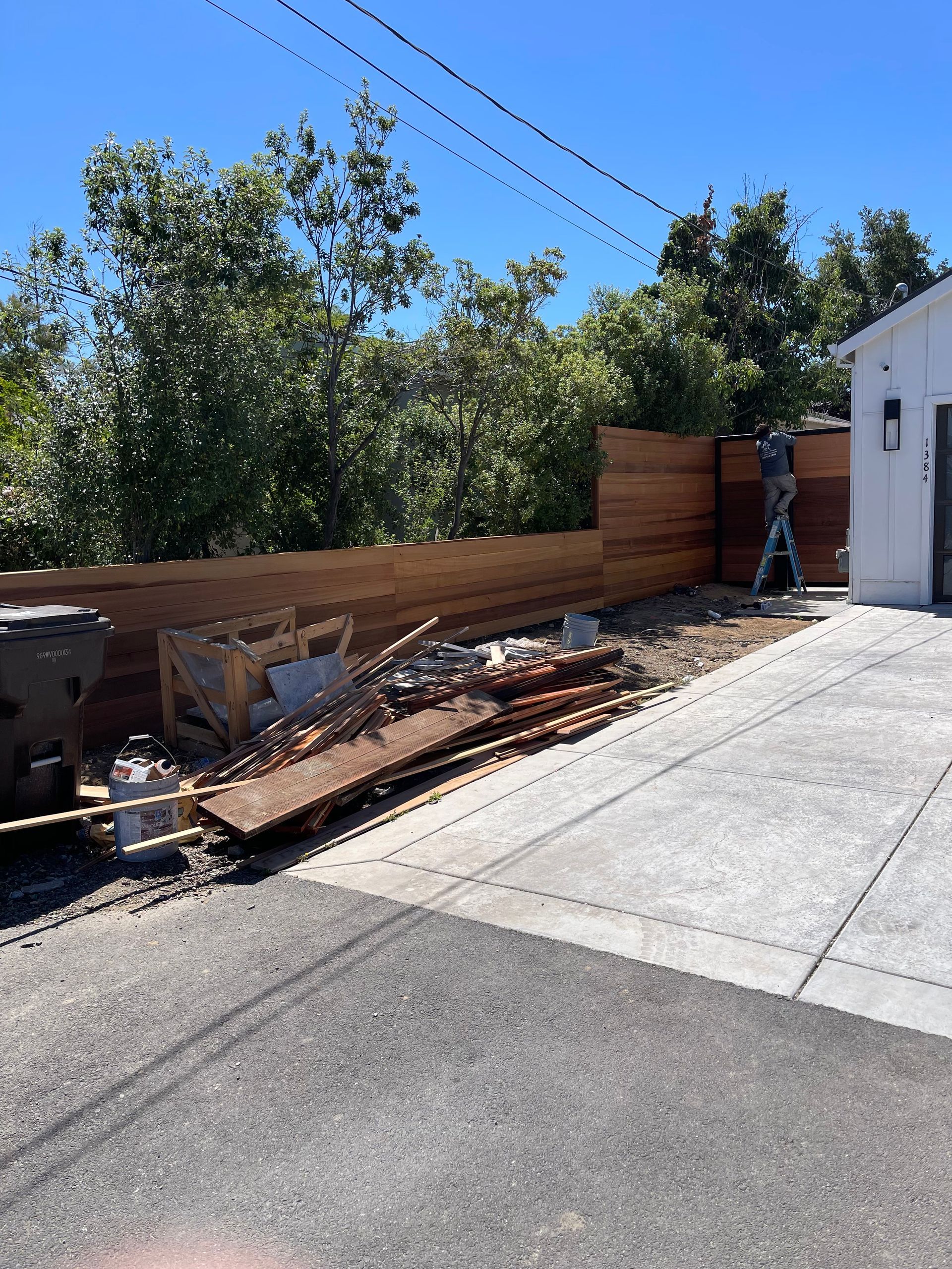 A person on a ladder installs a new horizontal wooden fence next to a house and a driveway with construction debris.