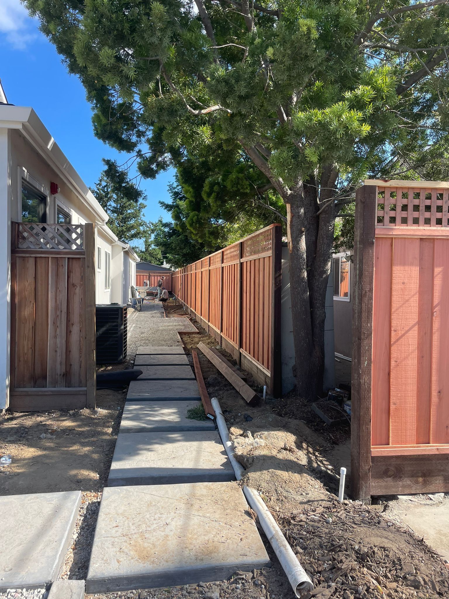 A narrow outdoor walkway featuring stone pavers leading between a house and a wooden fence, shaded by a large tree.