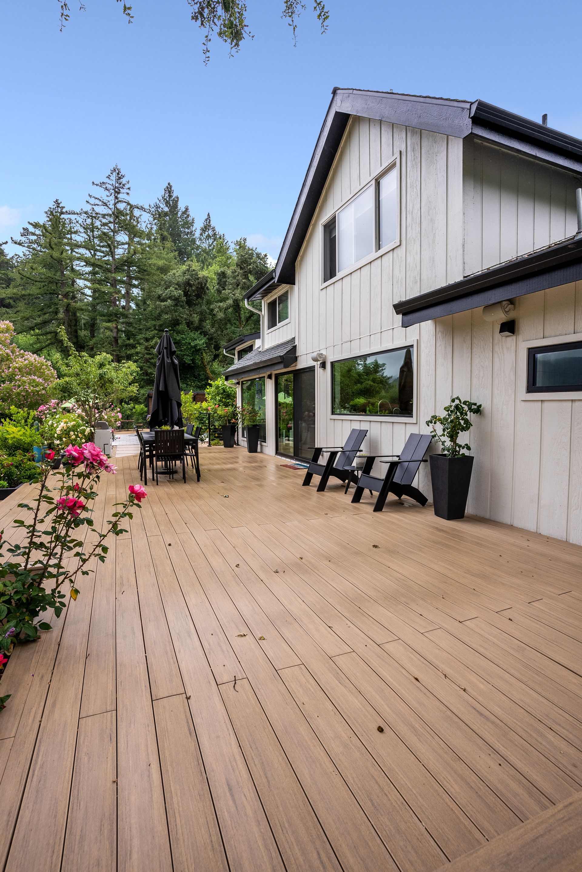 Wooden deck beside a white house with patio chairs, flower pots, and trees under a blue sky