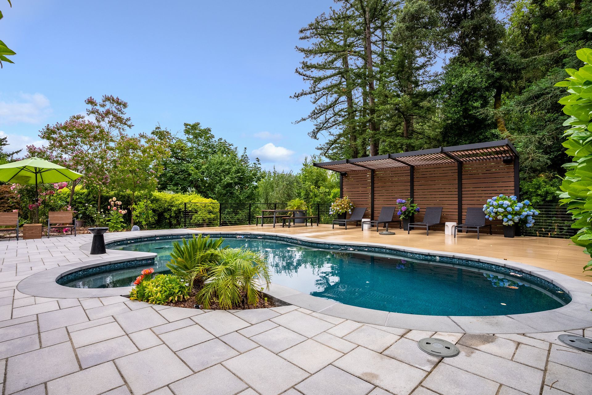 Backyard pool with stone patio, pergola seating, and lush green landscaping