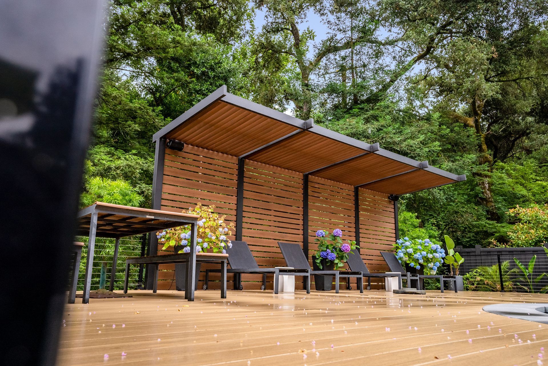 Outdoor wooden shelter with chairs and tables on a wet patio, surrounded by lush green trees