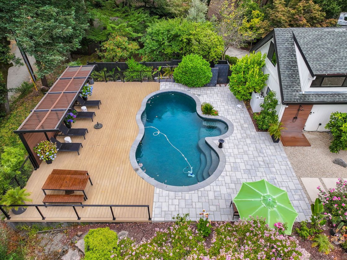 Aerial view of a backyard pool with lounge chairs, pergola seating, and green umbrellas surrounded by landscaping