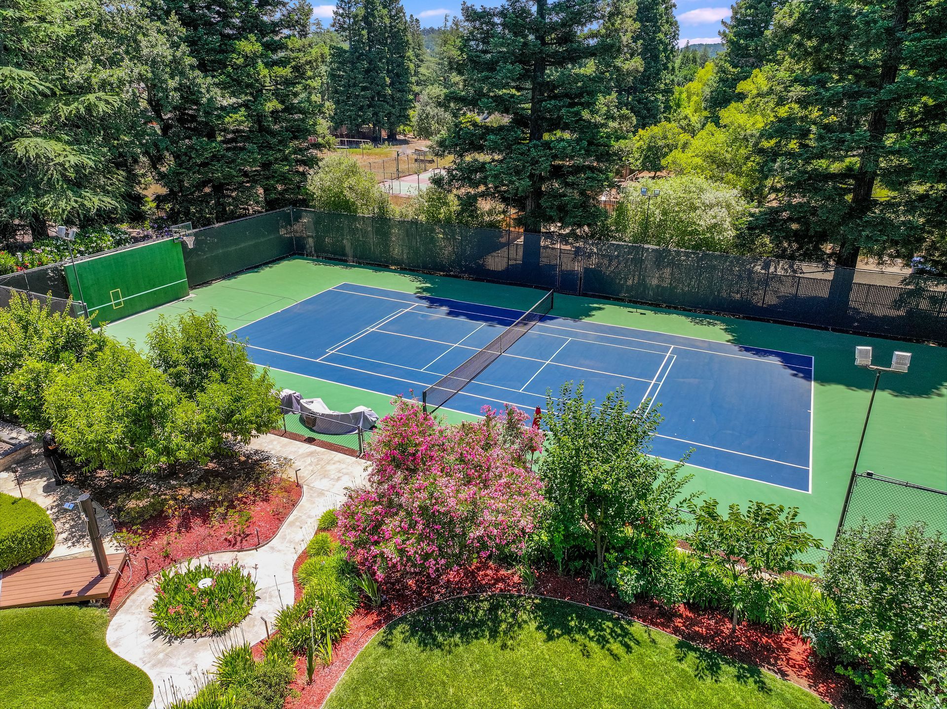 Outdoor blue tennis court bordered by green fencing, surrounded by trees and landscaped garden beds.