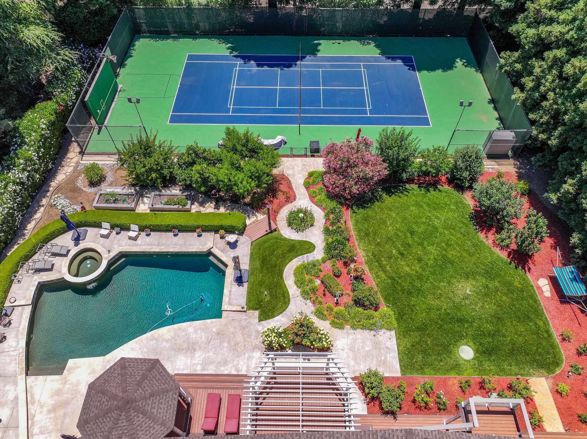 Aerial view of a backyard with a tennis court, pool, patio, and landscaped lawn.