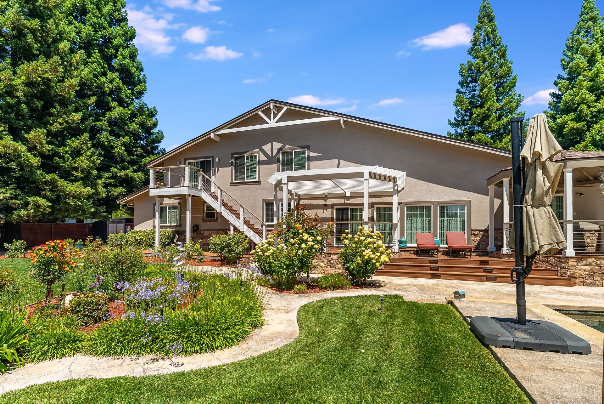 Landscaped backyard patio with a house, green lawn, flower beds, and a pergola under a blue sky