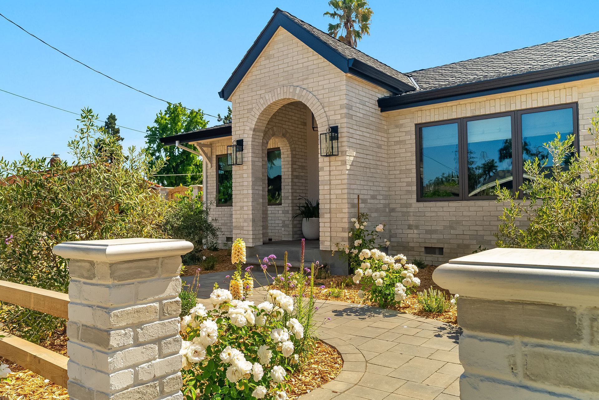 Stone house entrance with arched doorway, flower beds, and a paved front walkway under clear blue sky