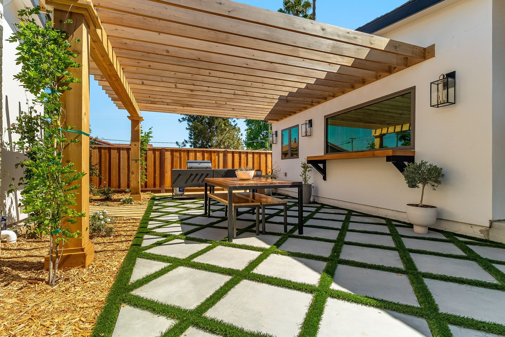 Modern patio with wooden pergola, outdoor seating, and geometric stone-and-grass paving by a white house