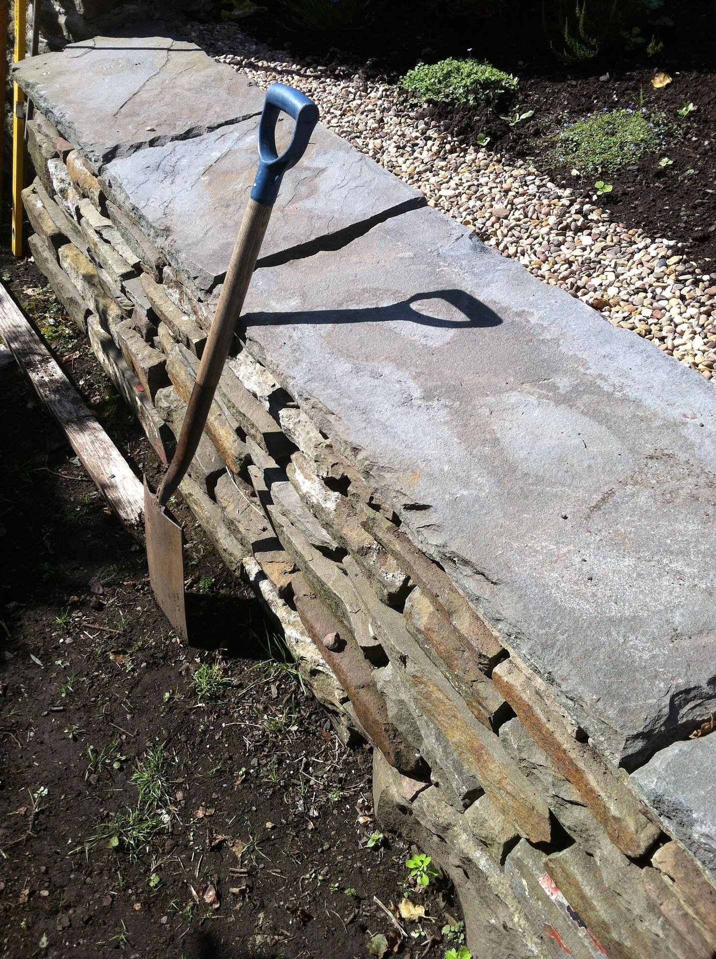 Shovel leaning against a low stone wall in a garden. Sunlight casts a shadow.