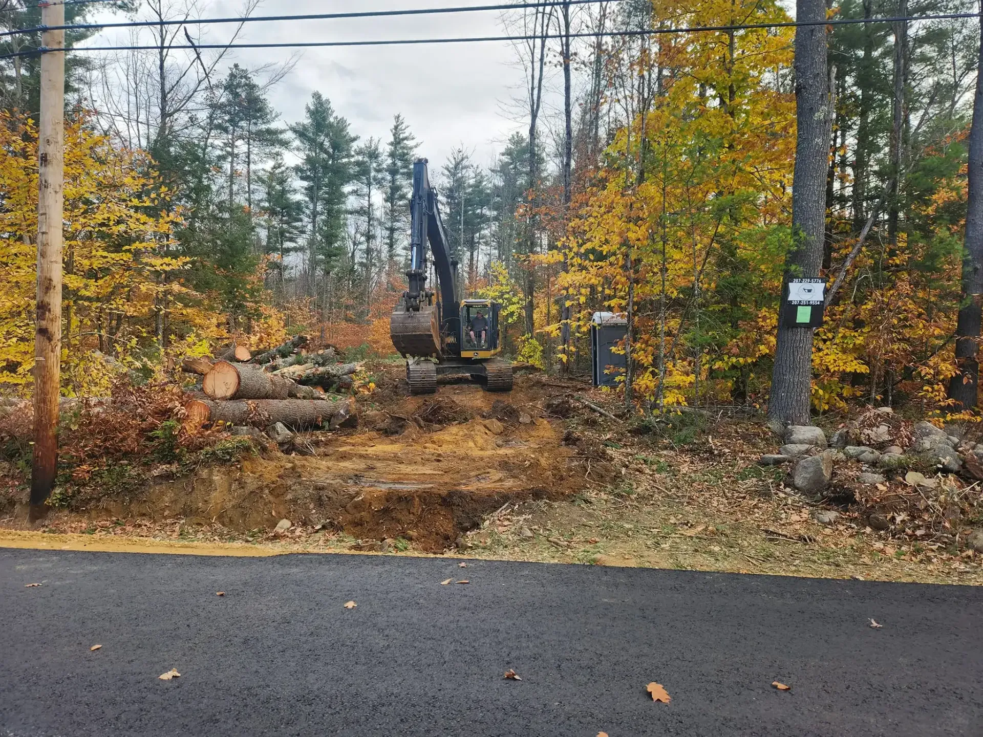 A large excavator is working in the woods near a road.