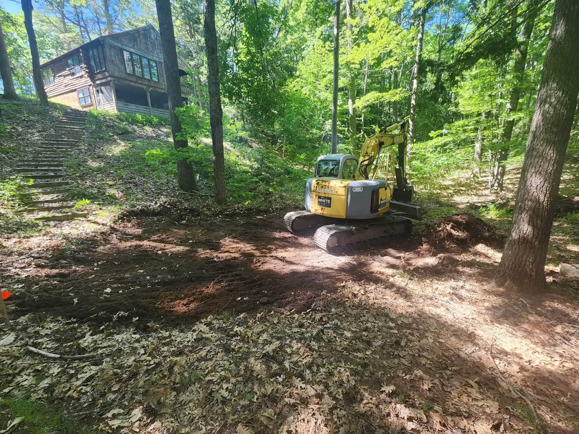 Mini excavator clearing brush in a wooded area near a house on a sunny day.