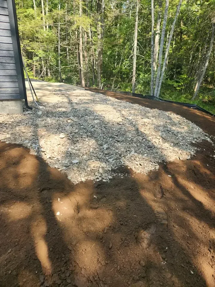 Gravel driveway under construction next to a dark gray house. Dirt and trees in background.