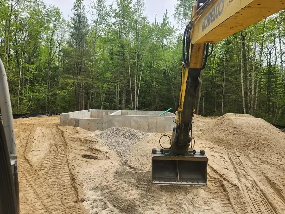 Excavator in front of a concrete foundation on a construction site with a forest background.