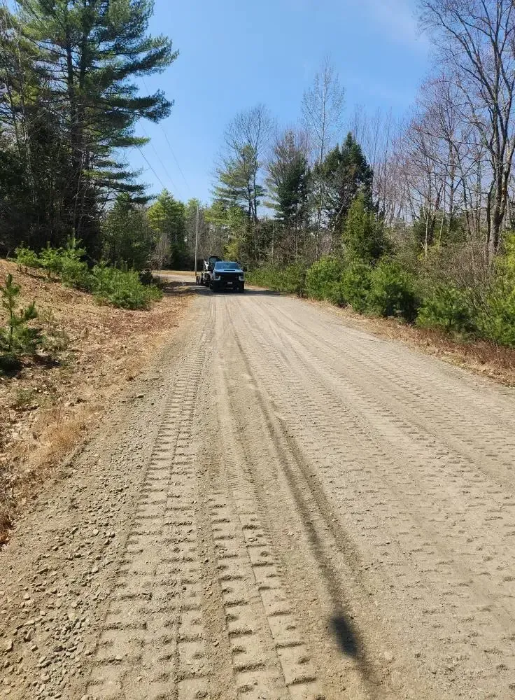 Gravel road leading to a dark vehicle, flanked by trees and greenery under a blue sky.