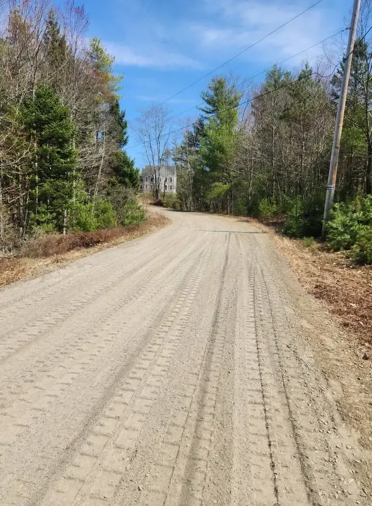 Dirt road through trees leads to a distant house under a blue sky.