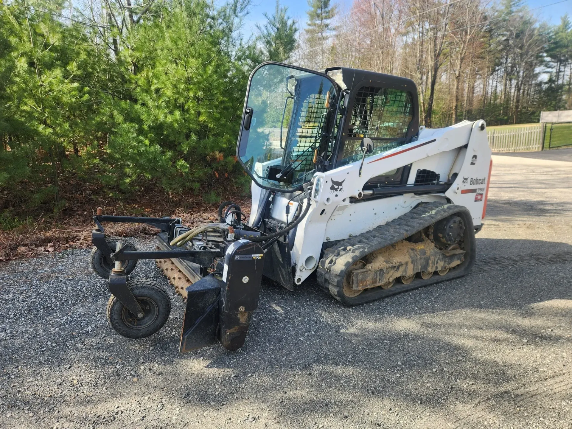 White Bobcat compact track loader with attached mower on gravel, near foliage.