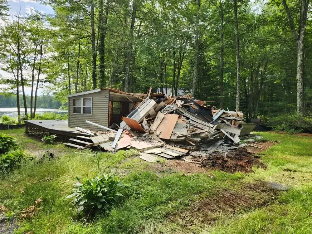 A partially demolished small house with debris pile in front, surrounded by green grass and trees.