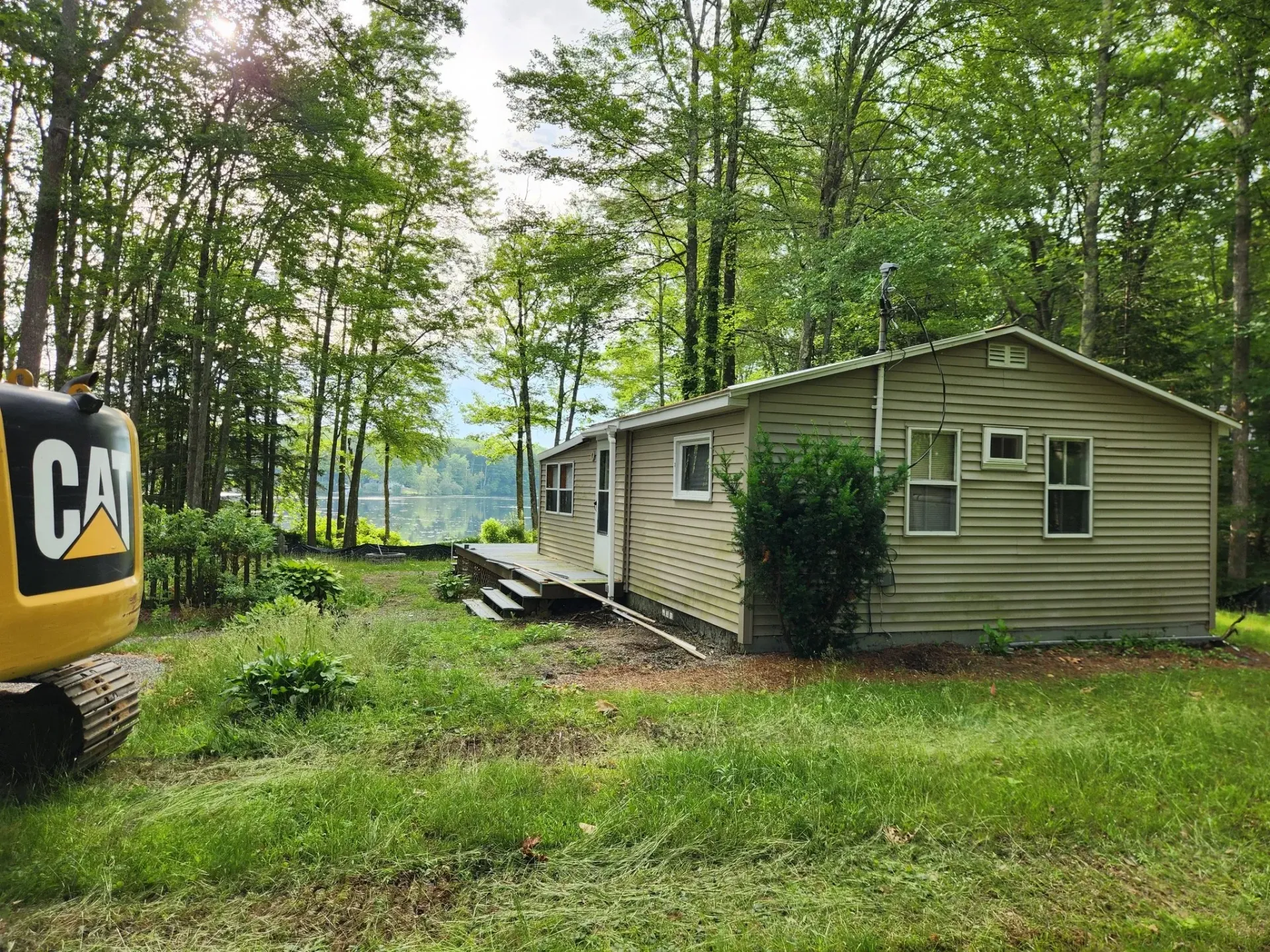 A small cabin with weathered siding, surrounded by overgrown grass and trees, and a parked CAT excavator.