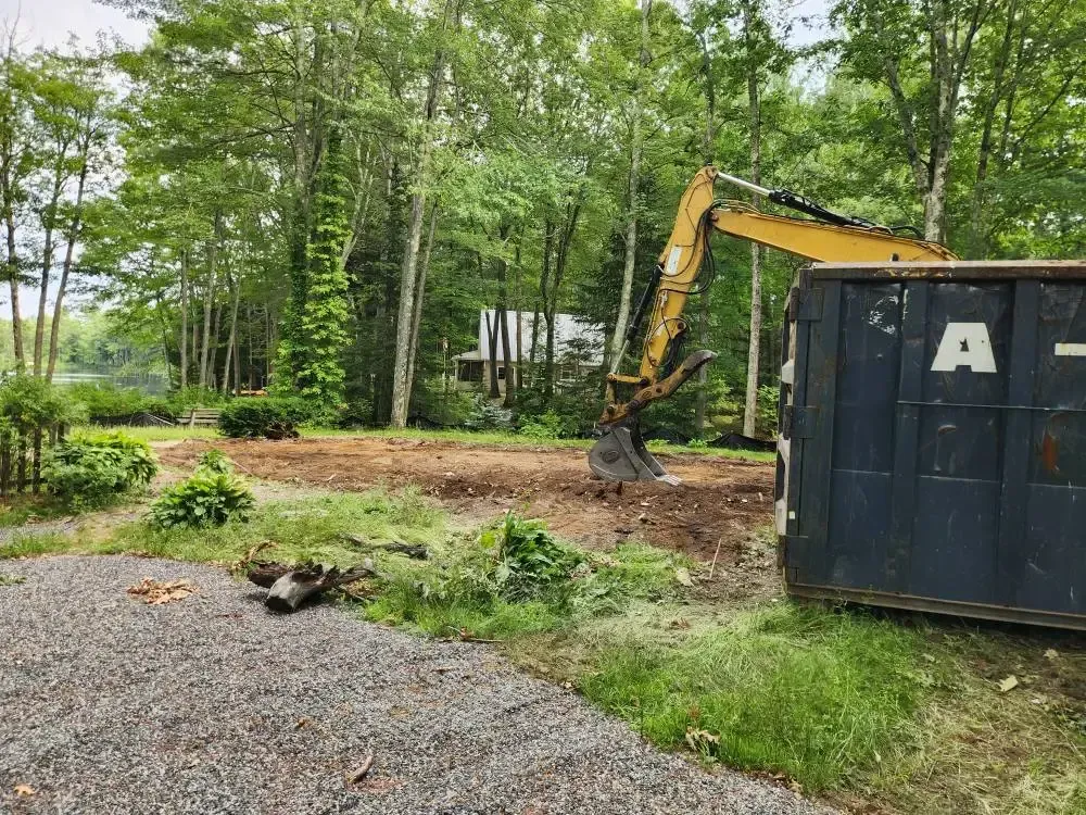 An excavator works on a cleared area next to a dark blue dumpster; trees and a house are in the background.