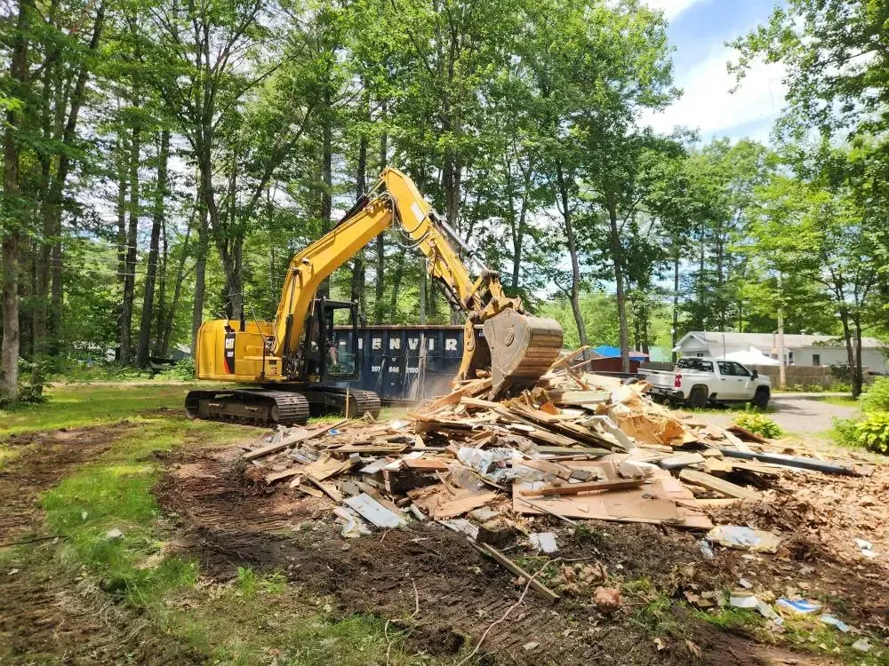 Yellow excavator loading debris into a blue dumpster in a wooded area. A white truck is parked nearby.