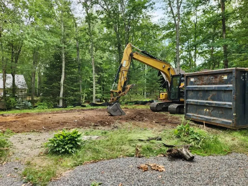 Yellow excavator on a dirt patch next to a dumpster in a wooded area.