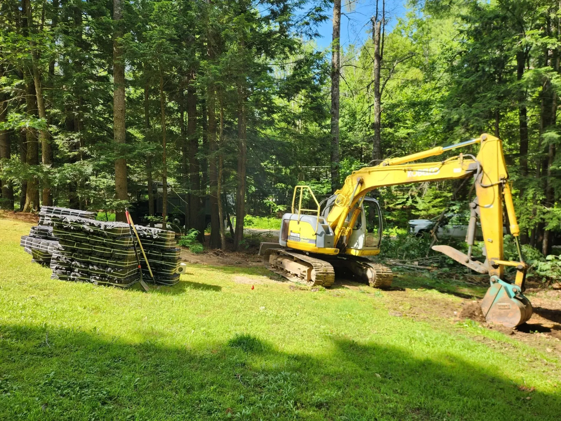 A yellow excavator is sitting in a grassy field next to a pile of logs.