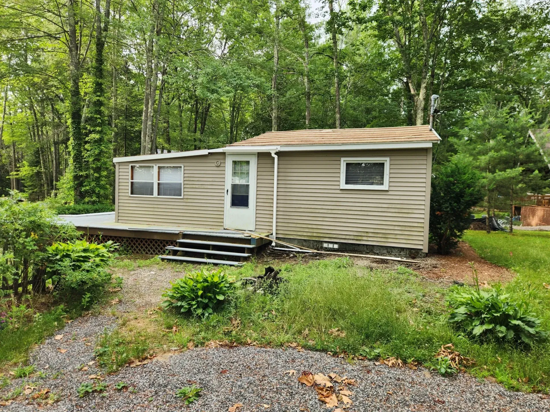 Small, light brown, single-story house with a small deck, surrounded by trees and grass.