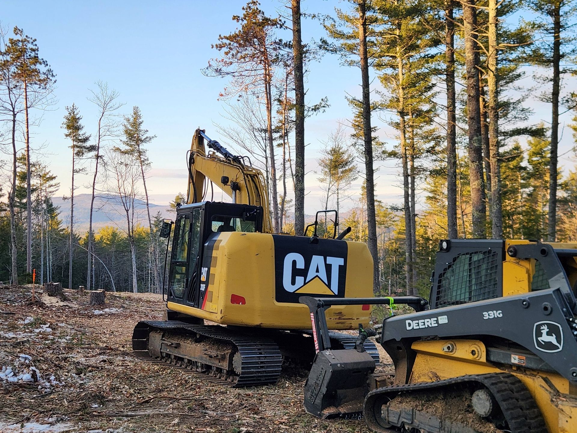 Yellow CAT excavator and John Deere skid steer parked on a forest clearing with scattered trees and wood chips.