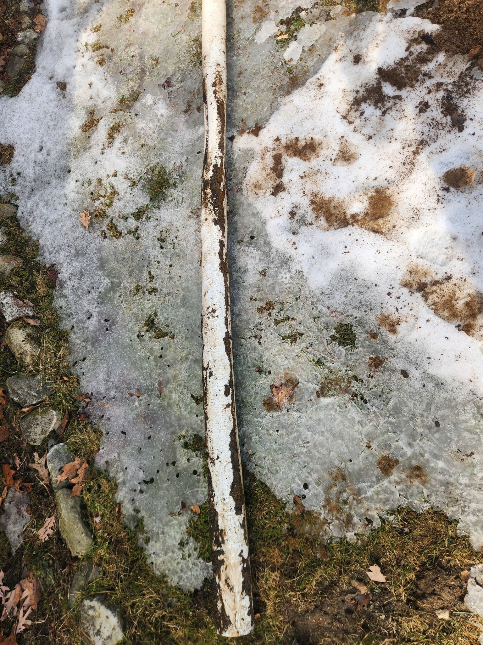 A weathered, white, cylindrical metal pole lying horizontally on ground covered in patches of snow and moss.